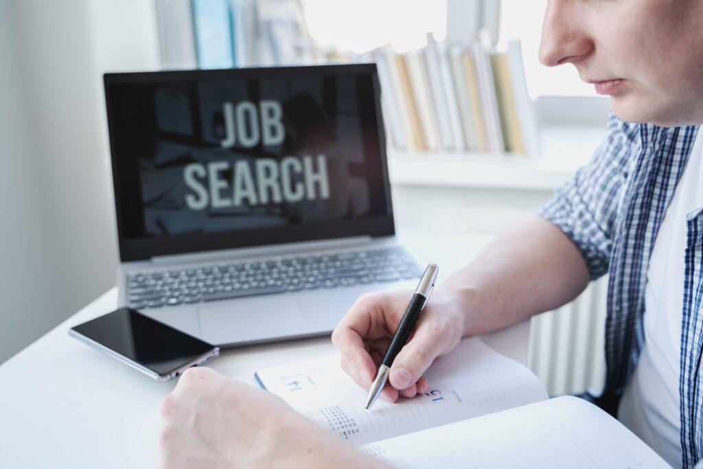 A young individual writing in a notebook while looking for job opportunities on his laptop